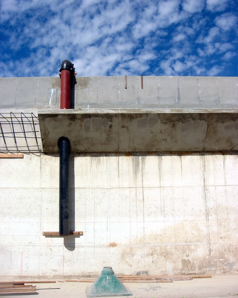 A concrete retaining wall under construction. There is a red and black plastic pipe that descends vertically through a cement ledge along the wall. Part of the ledge is incomplete with exposed rebar. On the ground are planks of wood and a green plastic cone of some kind. 