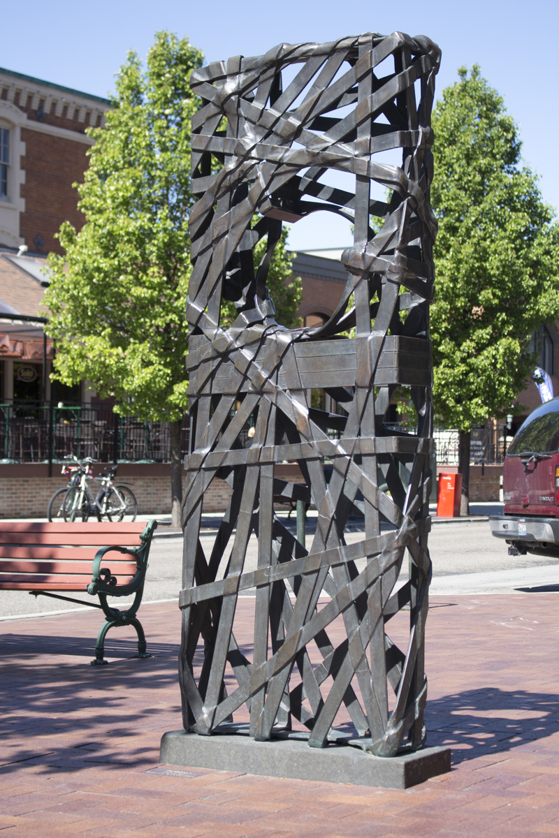 A seven foot tall rectangular hollow mass made from many loosely-woven overlapping bronze metal ribbons. At standing height is a circular opening, allowing a viewer to look through the sculpture. The sculpture is located on a brick-paved city sidewalk.