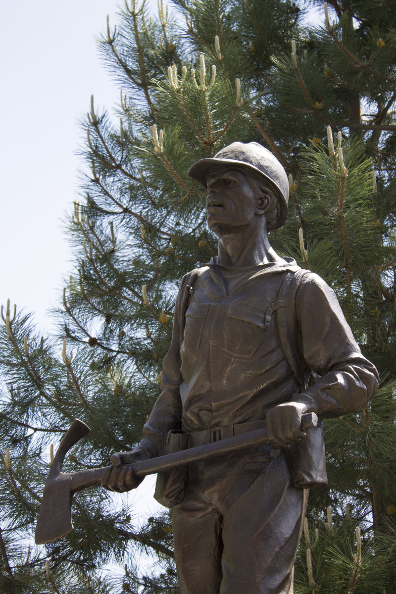 A bronze sculpture of a wildland firefighter wearing a helmet and holding a pulaski tool.
