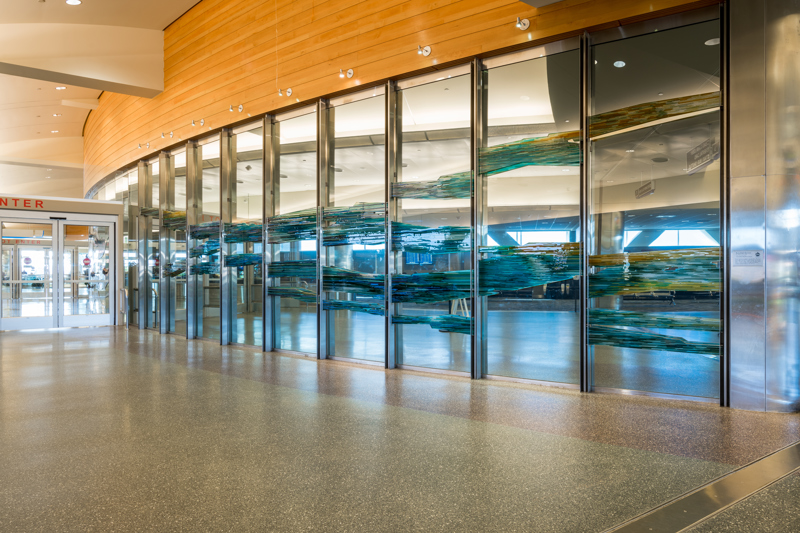 9 large glass panels which make up the wall separating the Boise Airport TSA check-in from the exit corridor. Each window is overlapped with thick geometric pieces of layered blue and yellow glass. From a distance, the composition of the colored glass resembles a horizon line along a mountainous landscape.