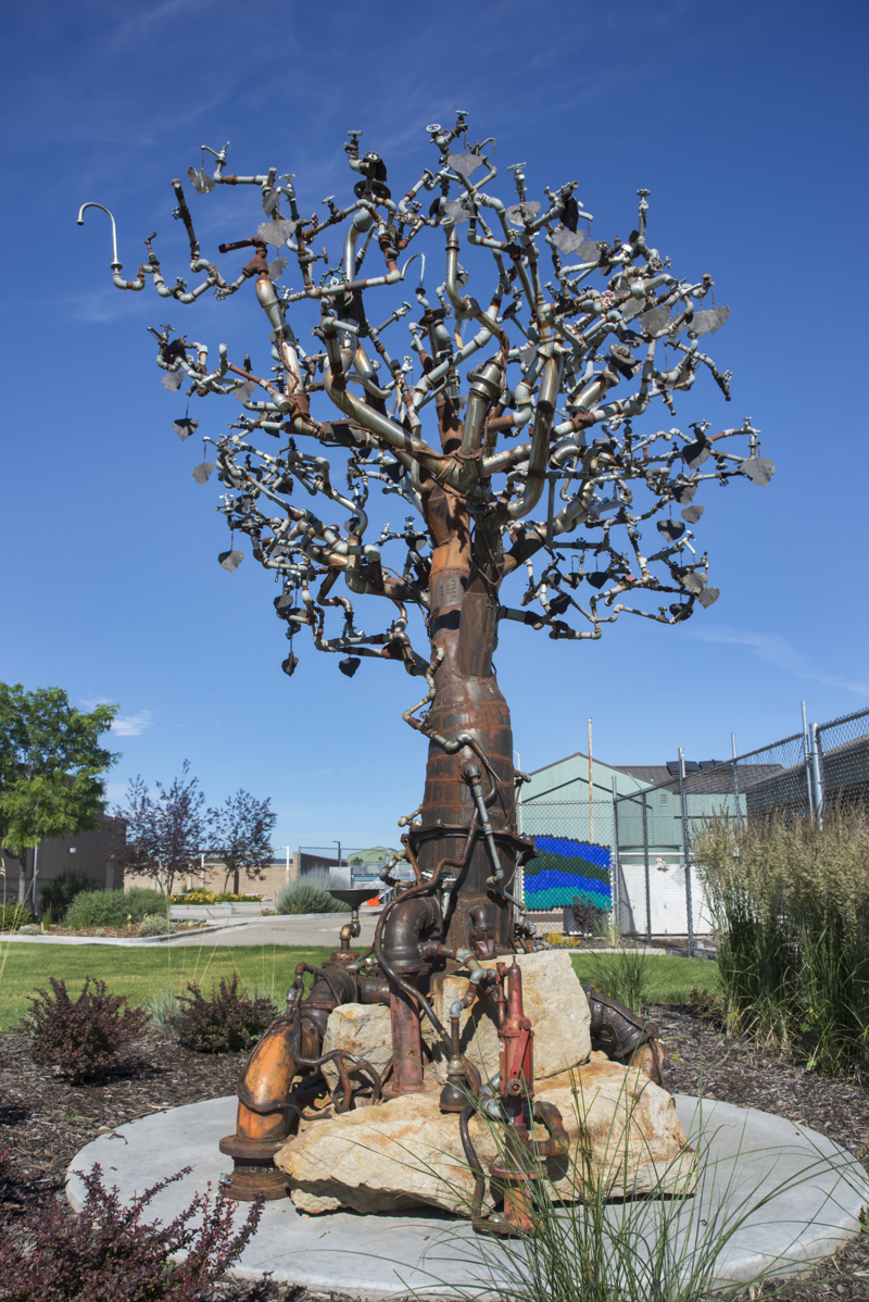 A large metal sculpture of a tree made from an assortment of metal plumbing artifacts including pipes, spickets, tubes, water meters, and fountains. The sculpture sits upon several large sandstones. Attached to its branches are over 150 bronze-cast leaves.