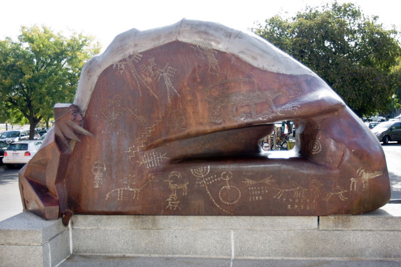 A weathered and stainless steel sculpture that depicts a Native American clinging to a rock formation that features petroglyphs. 