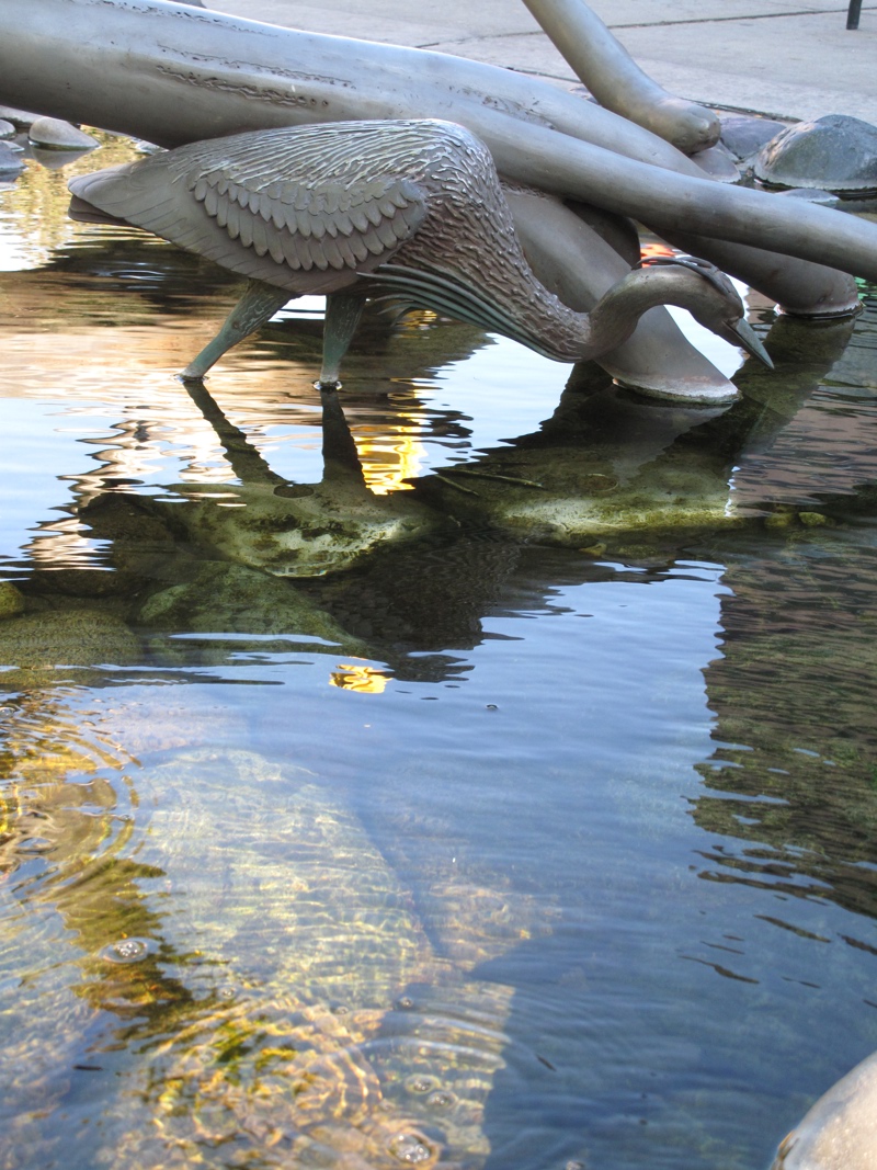 A water feature with an accompanying sculpture. Water flows as a small stream over smoothed river rocks of different colors beneath a stainless steel sculpture of three great blue herons on and around two fallen trees.