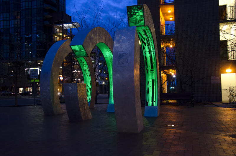 A sculpture made from brushed galvanized steel in the shape of three large semi-circular rings standing in a line parallel to one another. Lining the inner edges of the rings are inlays of clear acrylic plastic with screen prints depicting images and text referencing the history of Boise's Grove Street and the Boise Canal. At night the rings are internally illuminated with a green neon glow.