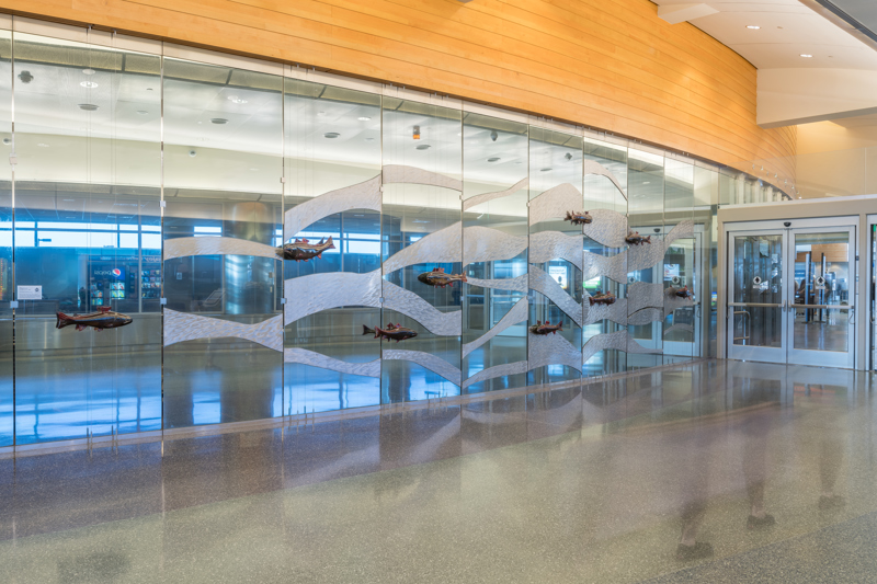 9 large glass panels which make up the wall separating the Boise Airport TSA check-in from the exit corridor. Over 8 of the windows are cut sheets of brushed stainless steel. From a distance, their composition resembles the waves of a river or stream. Overlapping several panels, suspended from tight cables, are 9 blown glass fish.