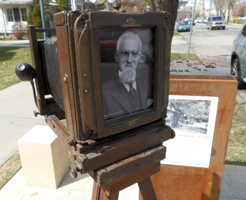 A life-size bronze sculpture of an antique tripod camera. In the viewing window of the camera is a portrait of a man (Jesus Urquides). In front of the camera is a stone pedestal for people to sit on as though having their picture taken. Next to the sculpture is a weathered steel pedestal topped with a model map of a community of houses (the Spanish Village). On the sides of the pedestal is signage describing the history of both Jesus Urquides and the history of the Spanish community in Boise. 