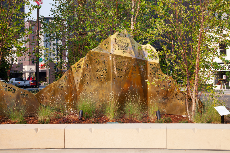 A geometric brass sculpture of mountain peaks topped with laser cut fabric patterns from various cultures. 