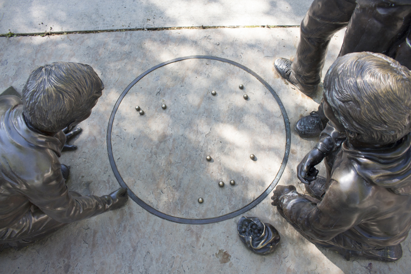 A life-sized bronze sculpture of three young children playing marbles on the sidewalk. 