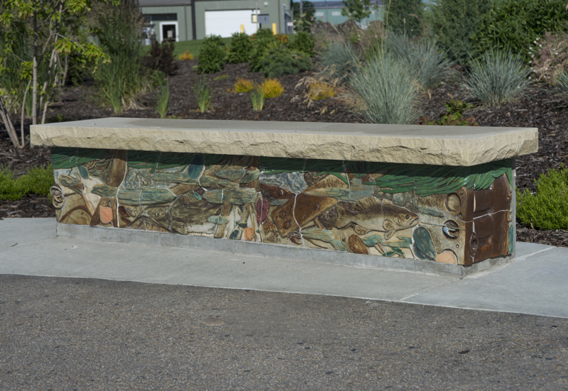 A bench made with a sandstone top and a base decorated with painted low bas-relief ceramic sculptures depicting a river bottom with rocks, litter, aquatic plants and animal life. Most notably is a fish.