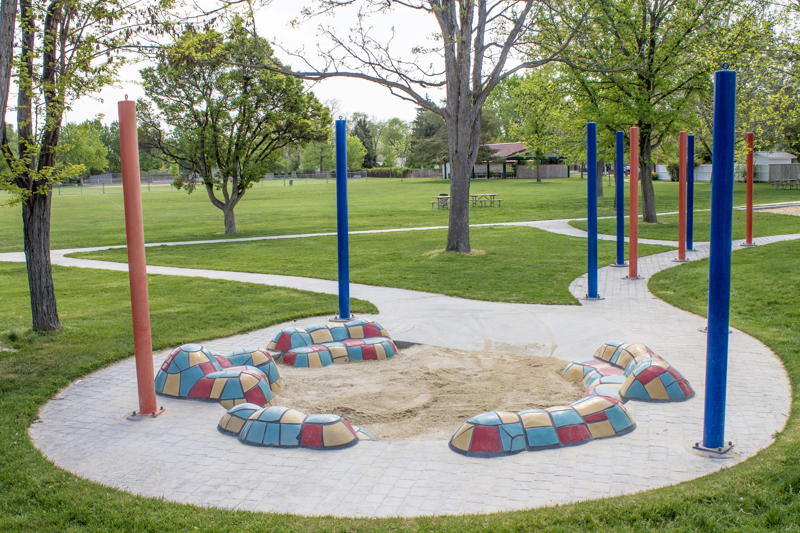 A small sand pit surrounded by sitting mounds covered by blue, yellow, and red ceramic tiles. The pit is encircled by brick and cement pathways leading off in different directions. Along the path are tall blue and red poles.
