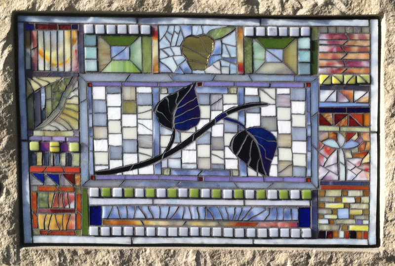 A closeup of one of the glass mosaics located on the side of the sandstone pedestal. The design has a day time theme with bright colors and warm tones being used.