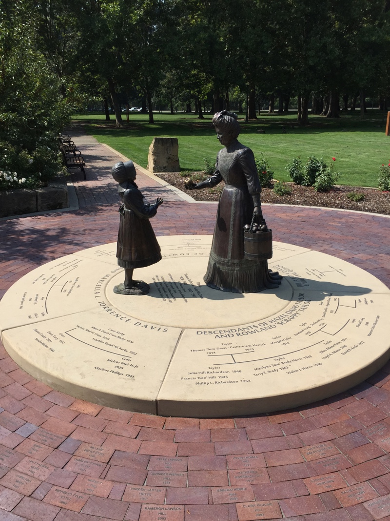 A life size bronze sculpture of a woman dressed in Victorian Era clothing (Julia Davis) holding a bucket of apples, and handing an apple to a young girl.