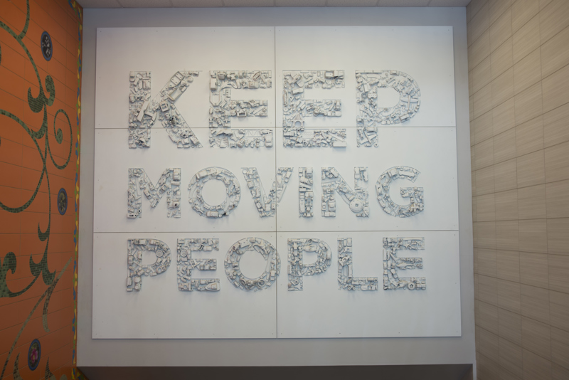 A large installation of six panels with the text, "Keep moving people". All text is filled with toys of various vehicles and modes of transportation that have been spray painted white. 