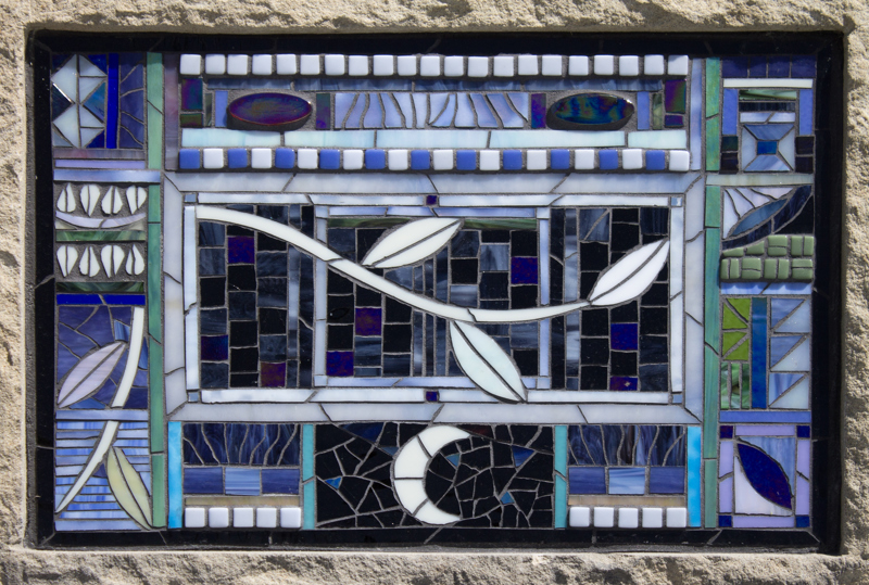 A closeup of one of the glass mosaics located on the side of the sandstone pedestal. The design has a night time theme with darker colors and cool tones being used.