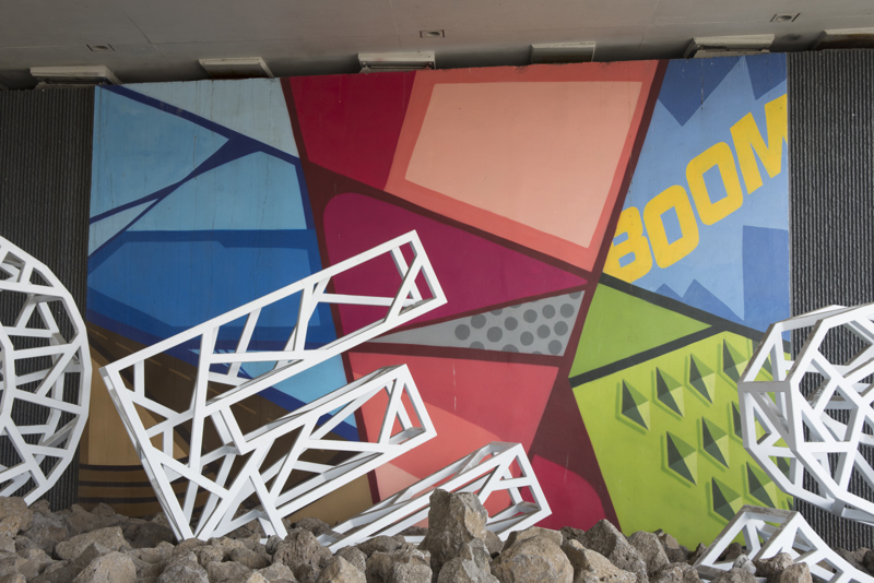 A mural painted in five parts on the wall of a highway overpass. All five sections are composed of geometric shapes with cartoon-like outlines and block colorations.