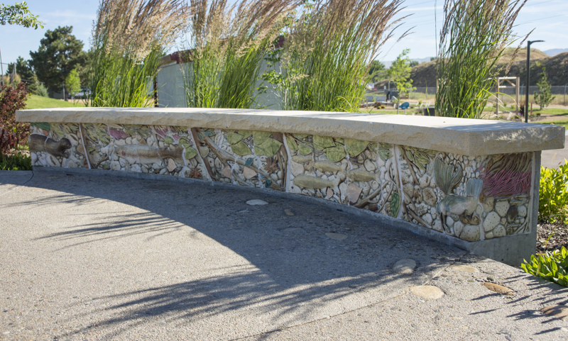 A curved bench made with a sandstone top and a base decorated with painted low bas-relief ceramic sculptures depicting a river bottom with an assortment of aquatic animals including a river otter, fish, and a kingfisher. 