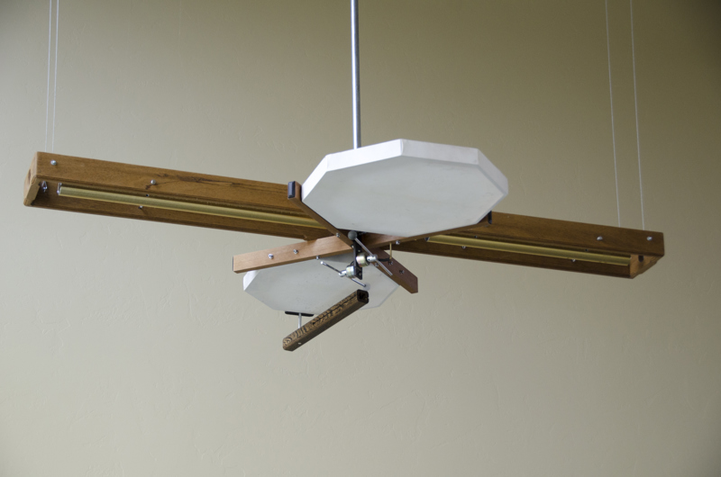 A kinetic sculpture made up of various percussion instruments including two stretched-leather drums, and two metal tube bells, all held together with a varnished wood structure. There are mallets that strike the instruments in a pattern and frequency that is determined by the direction and strength of the wind outdoors.  