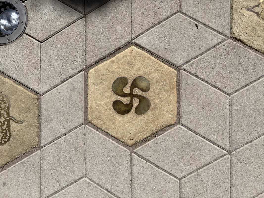 An image of a hexagonal sidewalk paver with a brass inlay of a Basque symbol.