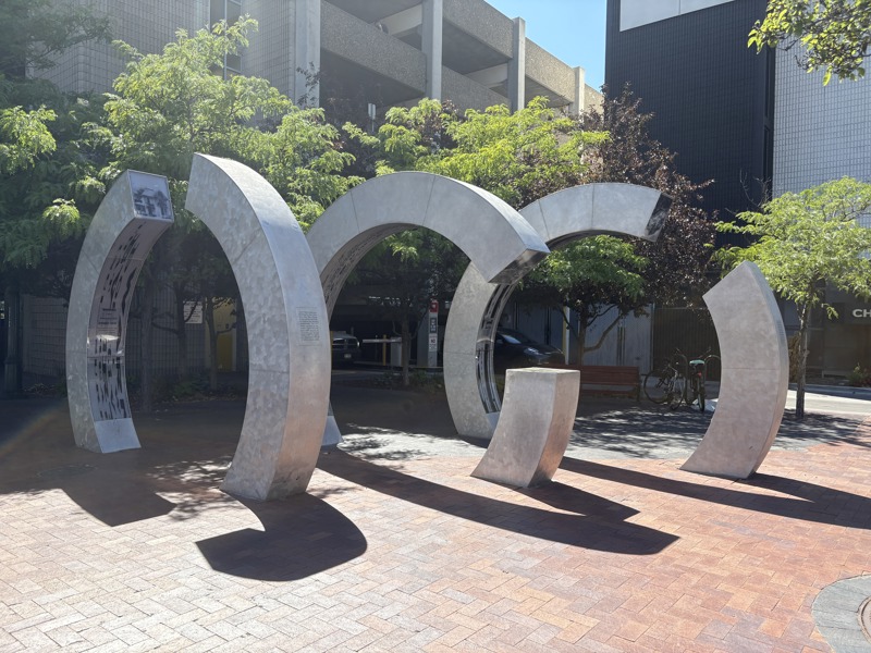 A sculpture made from brushed galvanized steel in the shape of three large semi-circular rings standing in a line parallel to one another. Lining the inner edges of the rings are inlays of clear acrylic plastic with screen prints depicting images and text referencing the history of Boise's Grove Street and the Boise Canal. 