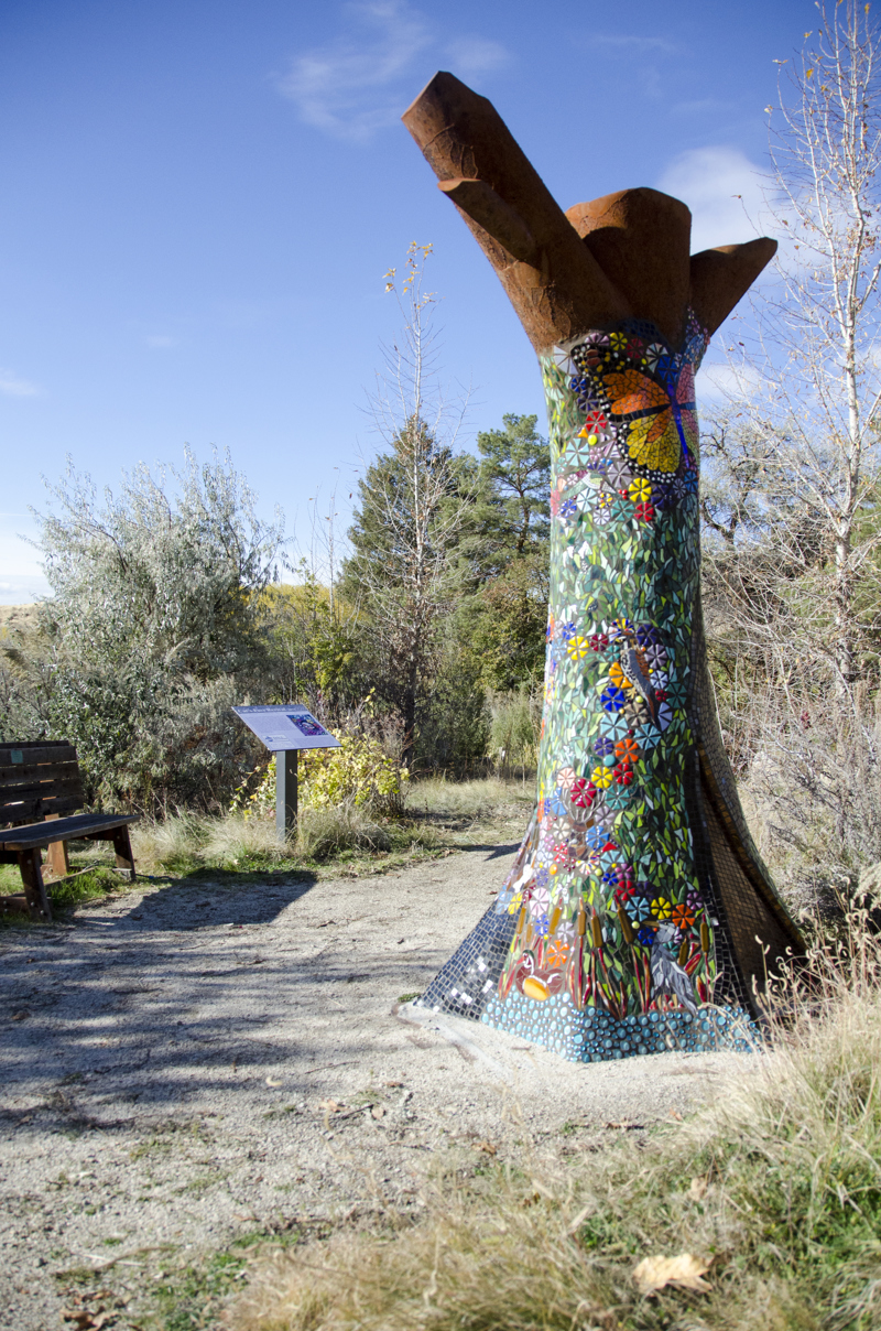 A life size sculpture of a dead standing hollow tree trunk. The sculpture is made of weathered steel and covered in glass tile mosaic depicting various lifeforms (i.e. birds, insects, and plants) that make use of dead or fallen trees in their habitat.