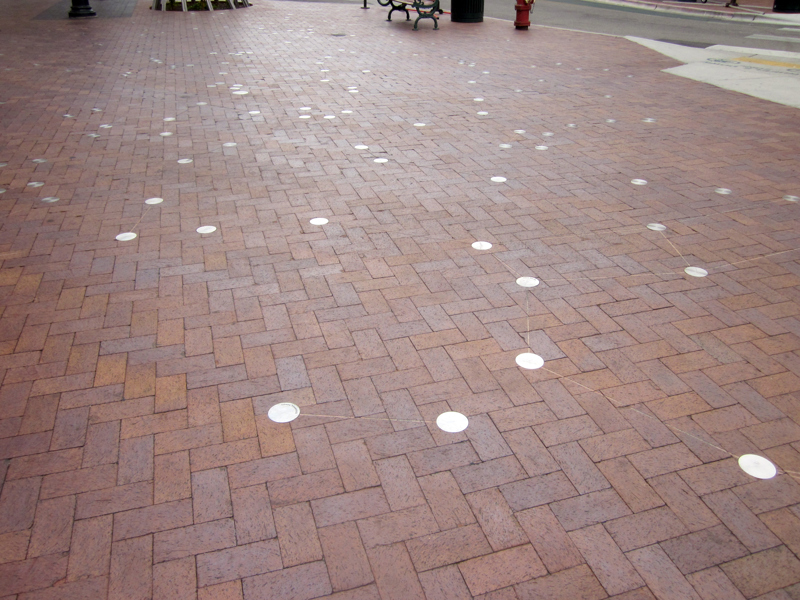 A collection of silver medallions inlaid into a cobble brick walkway. Lines are cut shallowly into the brick, connecting the medallions to form constellations.