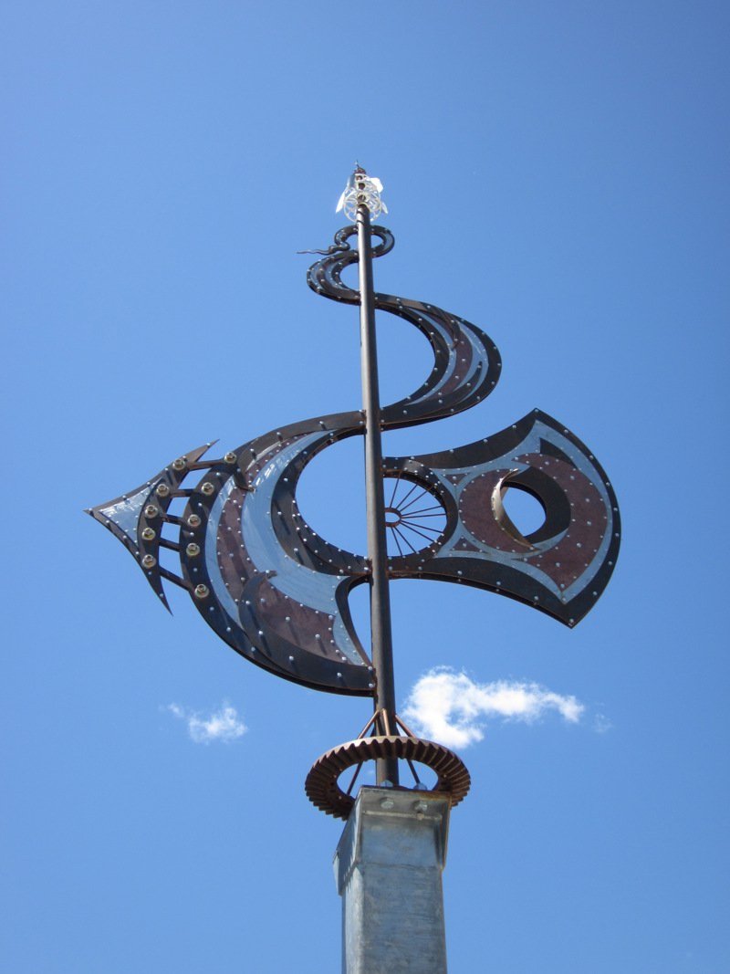 A tall metal sculpture resembling a weather vane. The base column is made from galvanized steel. The sculptural element at the top is made from a mixture of metals including Corten steel, stainless steel, and aluminum. It resembles a decorative letter S.