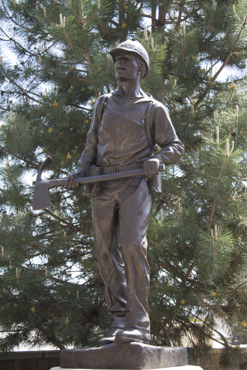 A bronze sculpture of a wildland firefighter wearing a helmet and holding a pulaski tool.