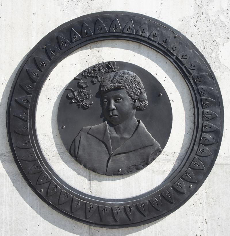 A low bas-relief bronze resin sculpture depicting a bust portrait of a young black woman. The sculpture is circular and is surrounded by a separate round ornate frame.
