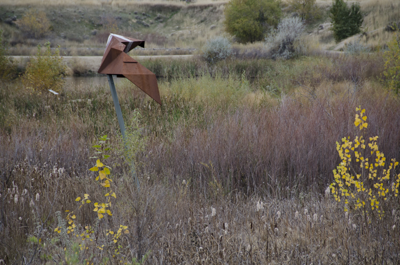 An abstracted geometric sculpture resembling a paper-folded origami bat in flight made of weathered steel, mounted onto a pole in the middle of a wetland.