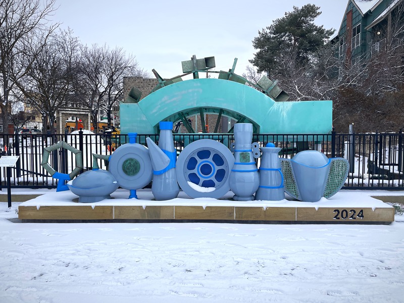 A sculpture depicting a lineup of 6 water clock vessels from different cultures in front of the waterwheel at Boise's C.W. Moore Park covered in snow.