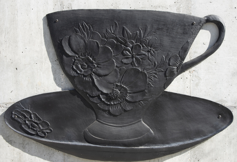 A low bas-relief bronze resin sculpture depicting a teacup on a saucer. The teacup is decorated with a flora design.