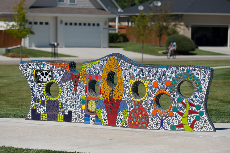 A short wall featuring a glass and tile mosaic surface. Along the wall are seven holes for peering through. The mosaic design showcases astronauts and aliens with the placement of the holes aligning with the figures' heads, creating a playful "face-in-hole" effect for looking through.