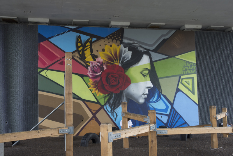 A mural painted in three parts on the wall of a highway overpass. On the left is the image of a yellow bird in flight, overlapping a leafy branch and a woman's bust. In the middle is an image of a woman in side profile with flowers in her hair. On the right is an image of a woman's upper back as she looks down. 