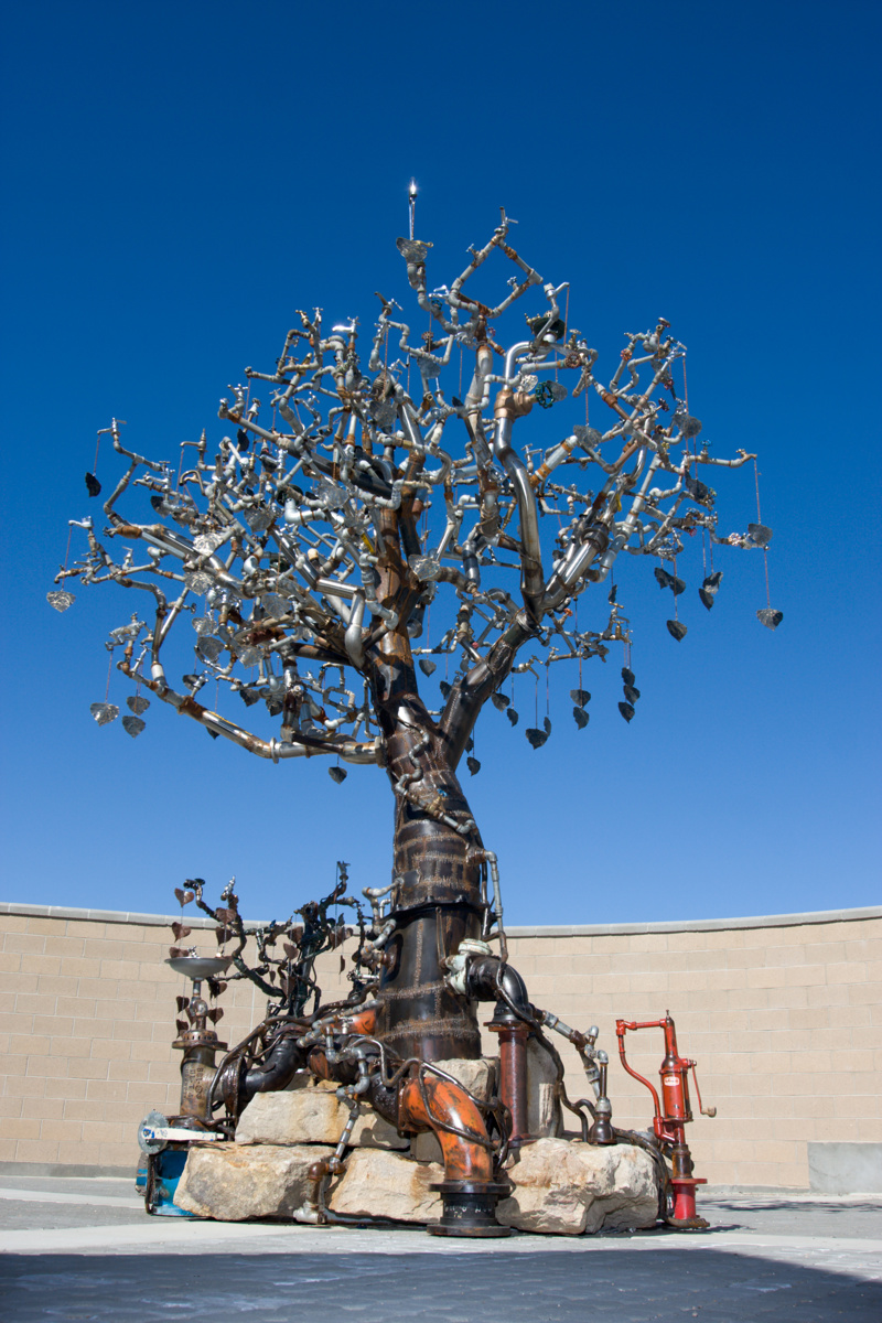 A large metal sculpture of a tree made from an assortment of metal plumbing artifacts including pipes, spickets, tubes, water meters, and fountains. The sculpture sits upon several large sandstones. Attached to its branches are over 150 bronze-cast leaves.