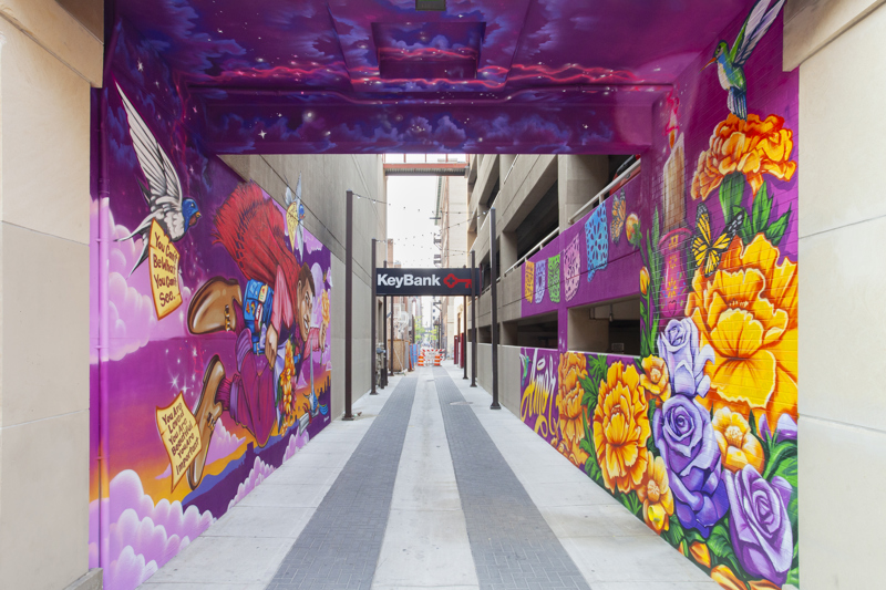 A painted mural that spans both sides of an alleyway. On one side is a dapperly dressed figure flying over the Boise skyline. They hold flowers and are surrounded by butterflies and swallows who carry notes of encouragement and affirmation. On the other side is an arrangement of flowers with butterflies, a hummingbird, a candle, and papel picado. On the far left is the word "Amore". 