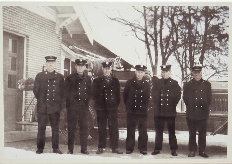 An image of 6 firefighters in uniform standing in front of a pumper truck in 1940.