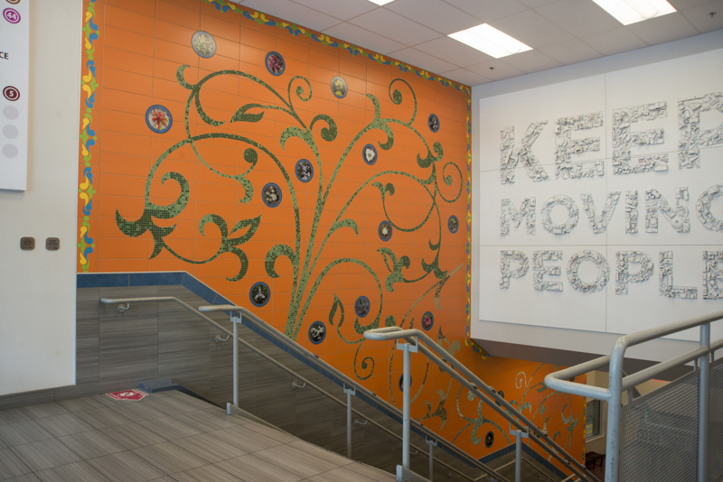 A mosaic wall installation made of ceramic tile. Made with green glass tile are leafy vines. Interspersed are tiles with printed images of local native flowers.