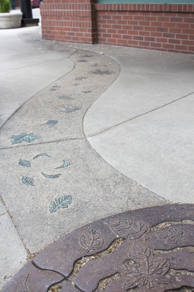A series of patinaed bronze leaves inlayed in pea gravel aggregate cement that curves within the regular cement of a city sidewalk, and turning around the corner of a red brick building.  The aggregate cement ends at the end of a rusty iron tree grate that has wavy lines resembling water ripples. In between the ripple are low-relief sculpted leaves that match the bronze leaves in the cement. 