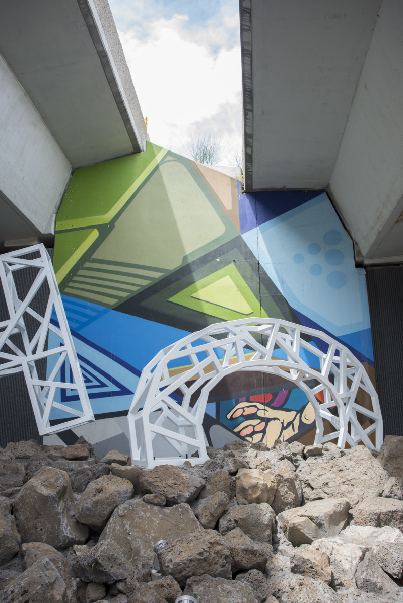 A mural painted in five parts on the wall of a highway overpass. All five sections are composed of geometric shapes with cartoon-like outlines and block colorations.