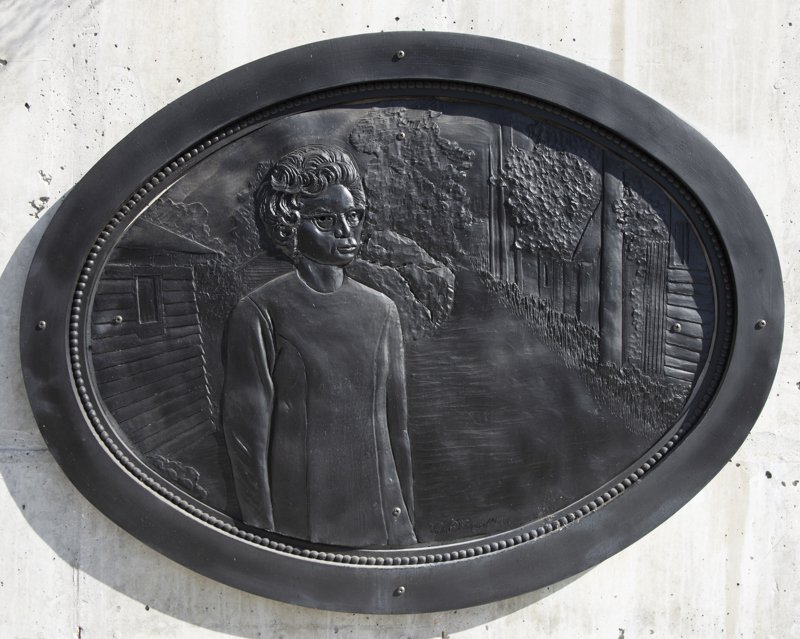 A low bas-relief bronze resin sculpture depicting a photo of a woman standing in front of an alley. The sculpture is an oblong oval shape and is surrounded by a separate ornate frame of matching shape.