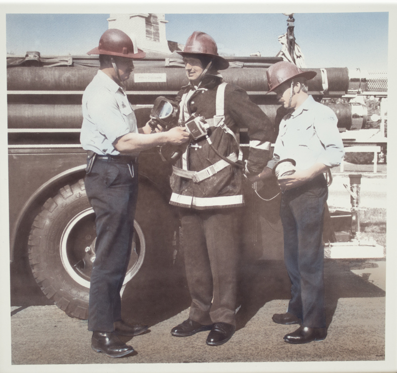 An image of three men standing in front of a vehicle. One is dressed in firefighting gear as he is being fitted for a mask.