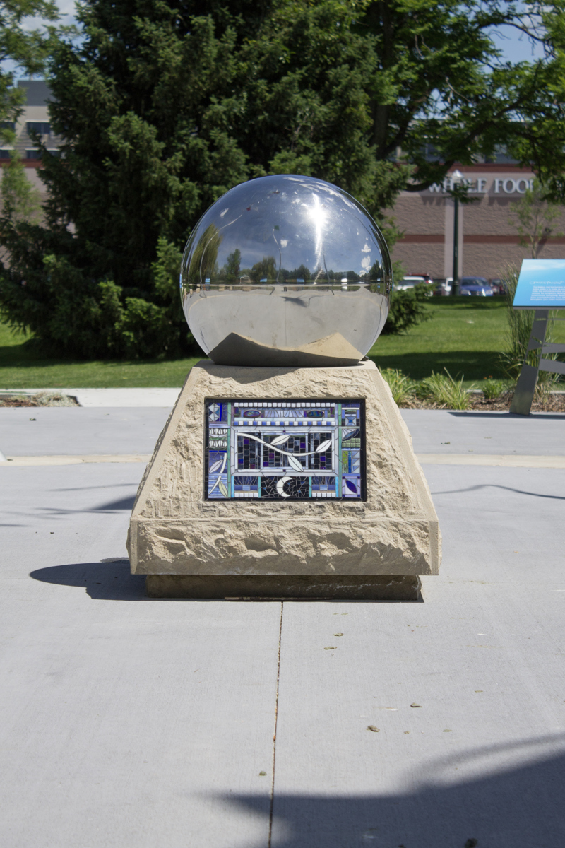 A 35" diameter steel gazing globe sitting atop of a sandstone pedestal. On the side of the sandstone pedestal is a glass tile mosaic.  