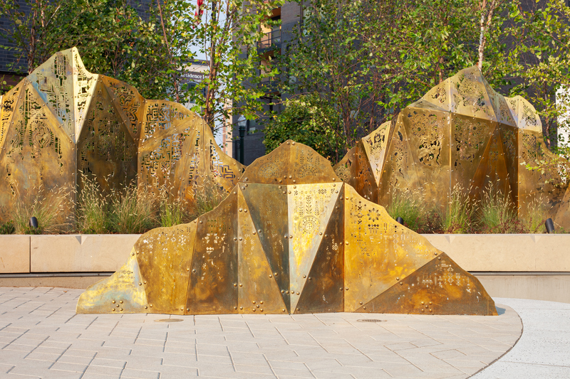 A geometric brass sculpture of mountain peaks topped with laser cut fabric patterns from various cultures. 