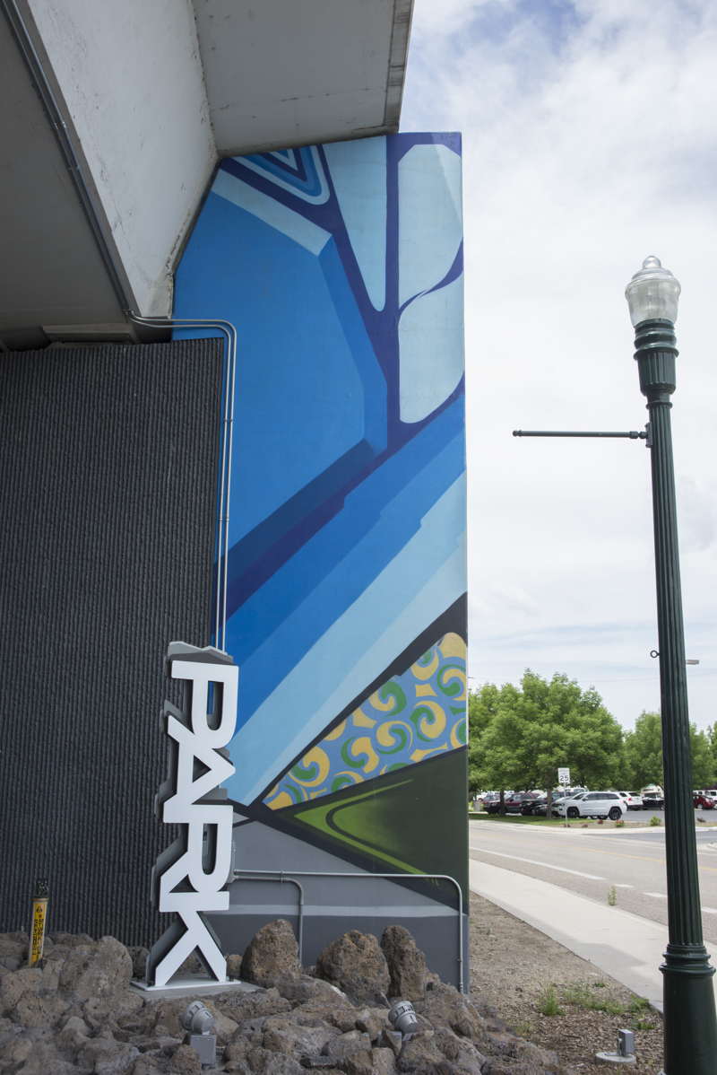 A mural painted in five parts on the wall of a highway overpass. All five sections are composed of geometric shapes with cartoon-like outlines and block colorations.