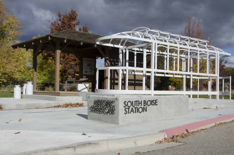 A historic streetcar station shelter made of wood. Outside are old metal milk containers painted white. In the interior are a suitcase and a basket made of metal painted white. Adjacent to the station is the metal skeletal frame of a streetcar painted white.