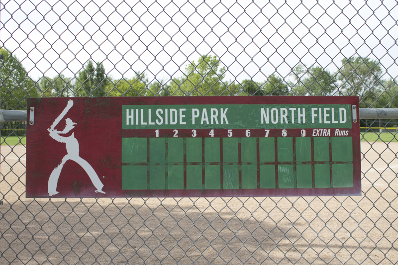 A baseball scoreboard attached to the field fence. On the left side of the scoreboard is an abstracted figure with a baseball bat. 