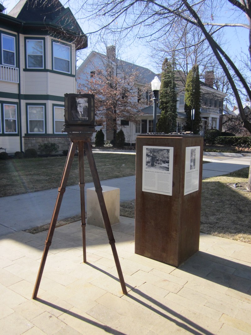 A life-size bronze sculpture of an antique tripod camera. In the viewing window of the camera is a portrait of a man (Jesus Urquides). In front of the camera is a stone pedestal for people to sit on as though having their picture taken. Next to the sculpture is a weathered steel pedestal topped with a model map of a community of houses (the Spanish Village). On the sides of the pedestal is signage describing the history of both Jesus Urquides and the history of the Spanish community in Boise. 