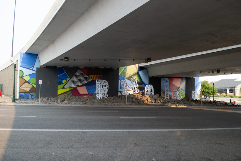 A mural painted in five parts on the wall of a highway overpass. All five sections are composed of geometric shapes with cartoon-like outlines and block colorations.