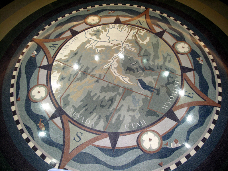 A terrazzo floor installation of a rose compass. In the middle of the compass is a map of the Northwest United States. Idaho is the only state in full view with Boise at the compass's center. The names of all the states that border Idaho (Washington, Oregon, California, Nevada, Utah, Wyoming, Montana) are written along the inner ring of the compass. On the outside of the compass, in the location of the intermediate directions, are four syringa flowers. Encircling the entire compass are fish in a