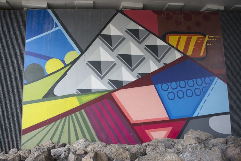 A mural painted in five parts on the wall of a highway overpass. All five sections are composed of geometric shapes with cartoon-like outlines and block colorations.